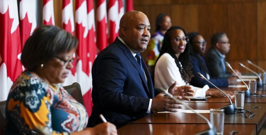 Wide shot of the Canadian Congress of Black Politicians founding executive panel seated behind a desk at a press conference on Parliament Hill with microphones and Canadian flags.