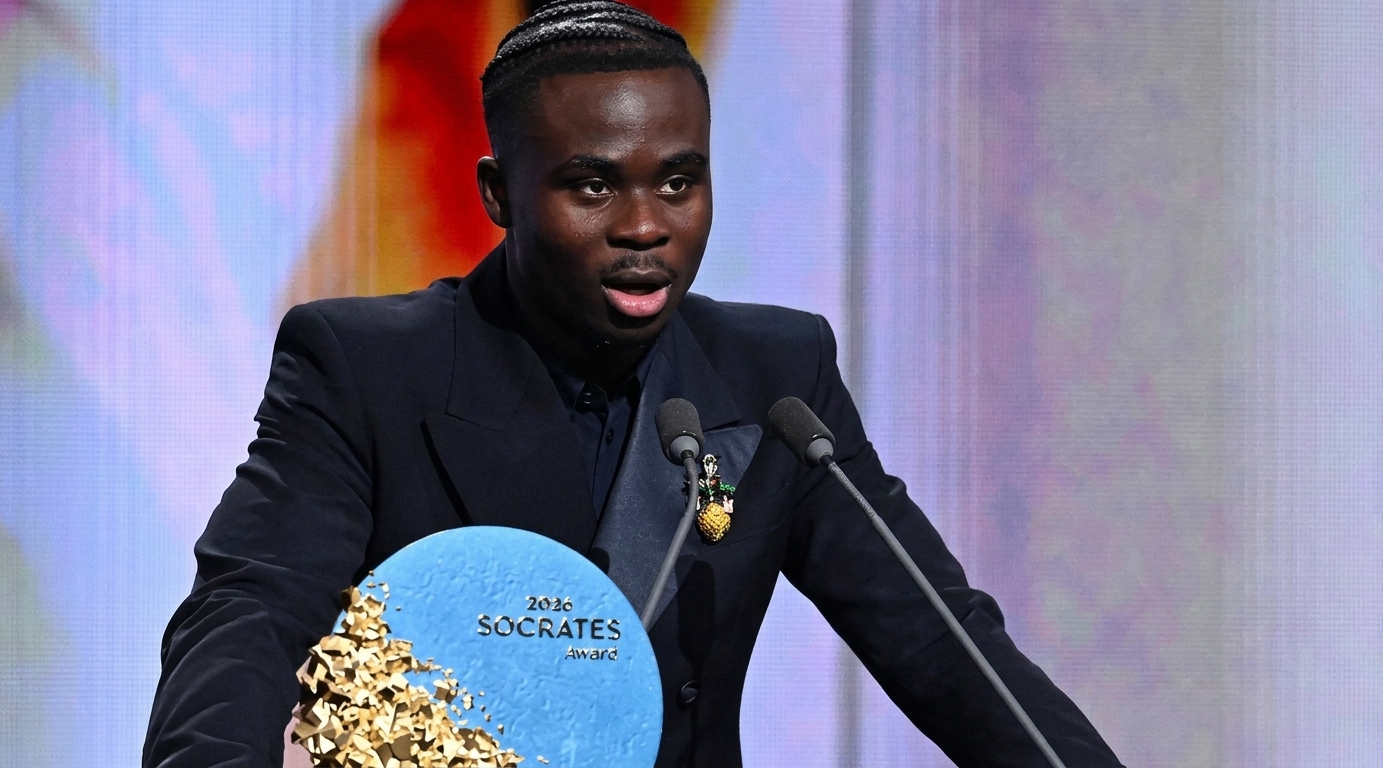 A crisp, close-up shot of Bukayo Saka in a sharp navy tuxedo speaking passionately into microphones at the Ballon d'Or gala podium.