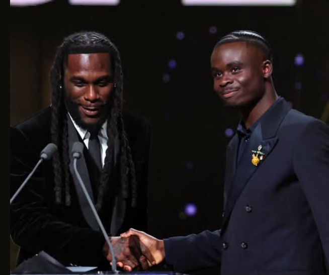 Nigerian music icon Burna Boy smiling as he hands over the bright blue and gold Sócrates Award trophy to a beaming Bukayo Saka on the main stage.