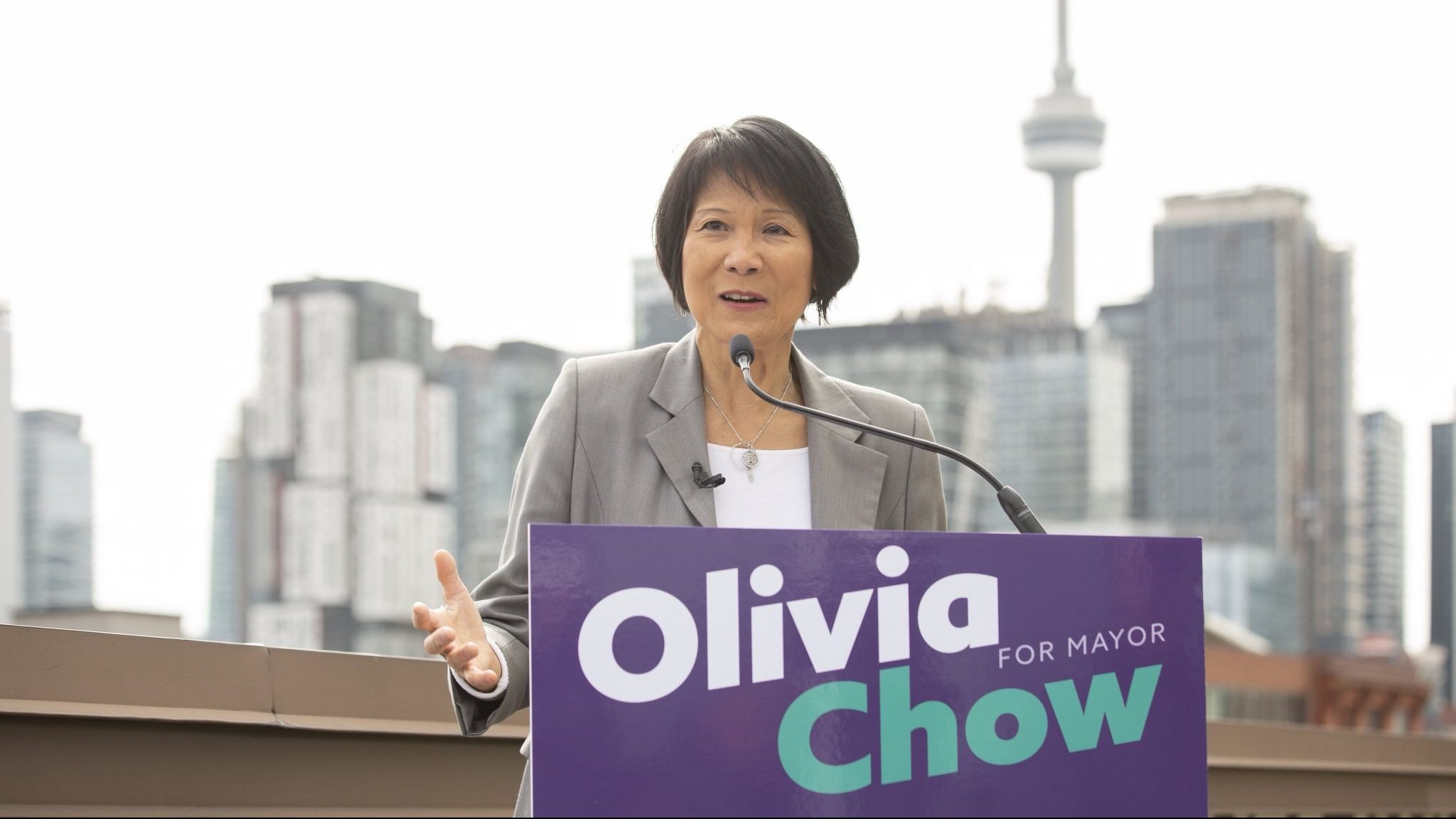 A portrait of Toronto Mayor Olivia Chow with short black hair, smiling warmly while wearing a bright yellow blazer over a white shirt and a circular silver pendant necklace. The background is a softly blurred outdoor transit vehicle.